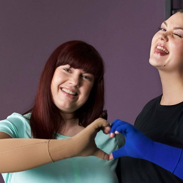Auf dem Bild stehen zwei lachende Frauen nebeneinander und formen mit ihren Händen ein Herz. Sie tragen beide eine VenoTrain curaflow Armversorgung von Bauerfeind, welche zur gezielten Ödemtherapie dient.
