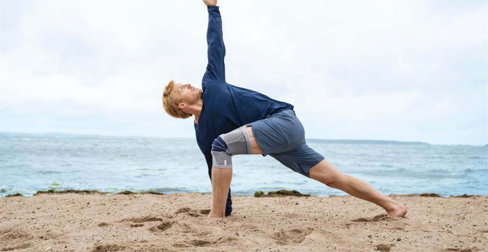 Das Bild zeigt einen Mann in Yoga-Pose am Strand. Am rechten Knie trägt er eine GenuTrain Bandage zur Unterstützung. Regelmäßige Knieübungen können Schmerzen lindern.