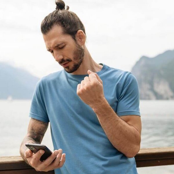 Das Bild zeigt einen Mann mit Smartphone, der an dem Ufer eines Bergsees steht. Er hält den rechten Arm mit schmerzenden Handgelenk hoch.