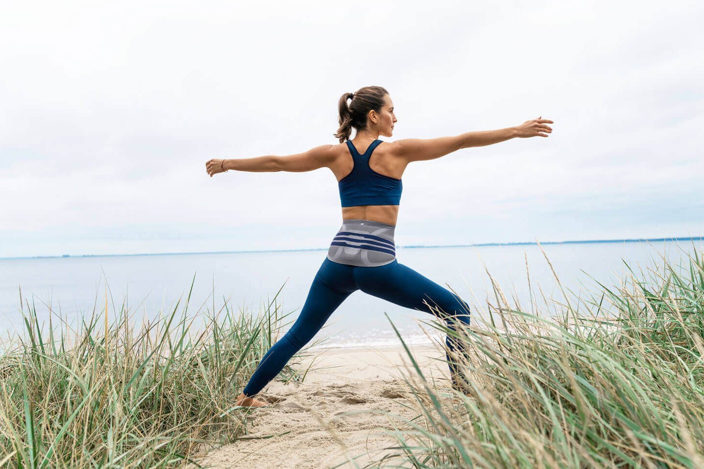 Das Bild zeigt eine junge Frau, die am Strand eine Yoga-Pose ausführt und dabei eine LumboTrain Lady Bandage zur Linderung von Rückenschmerzen trägt.