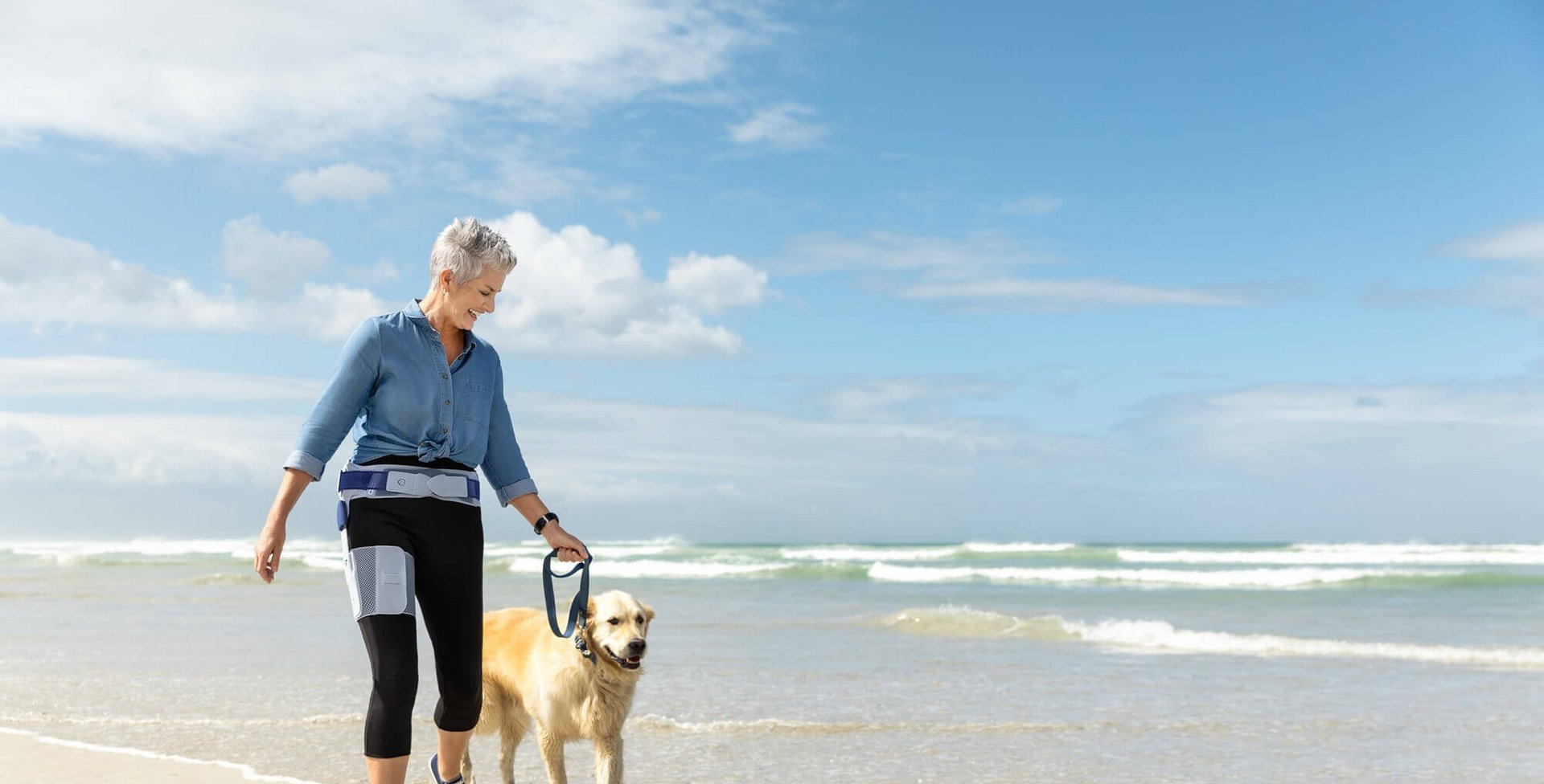 Das Bild zeigt eine ältere Frau beim Strandspaziergang mit ihrem Hund. Die Frau trägt eine CoxaTrain Orthese, die speziell für Hüftarthrose-Patienten entwickelt wurde.