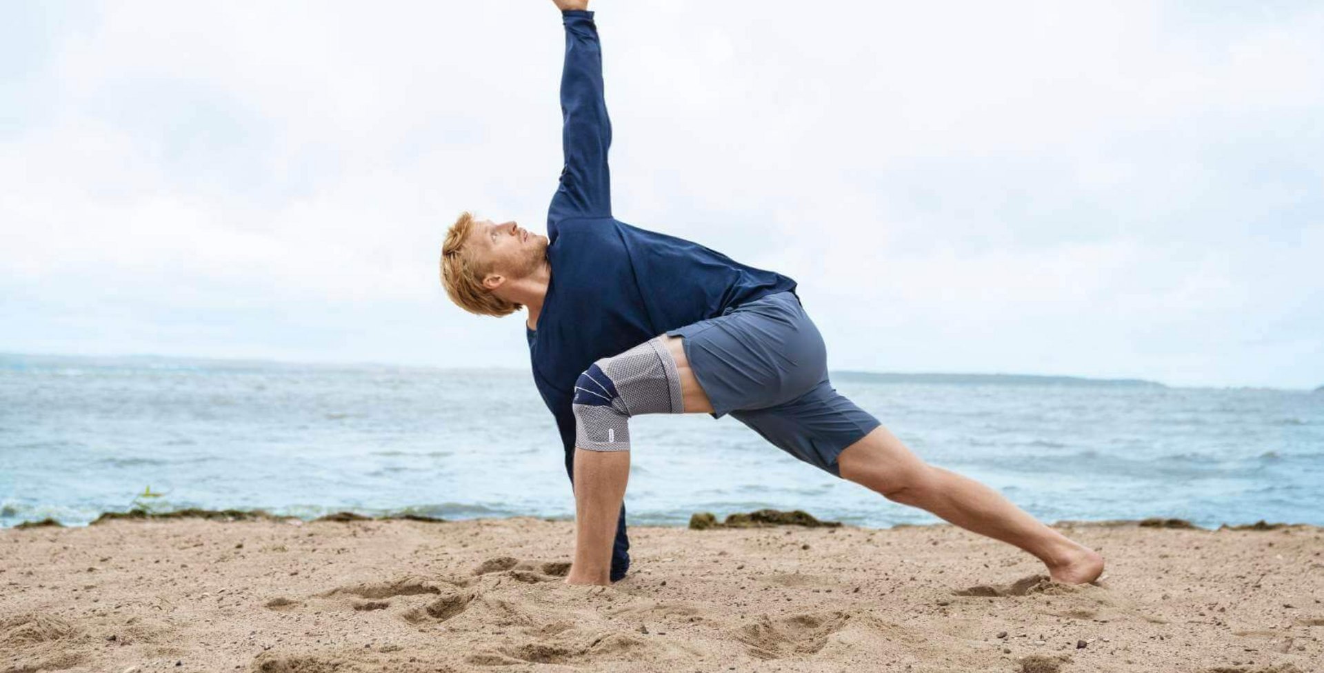 Das Bild zeigt einen Mann in Yoga-Pose am Strand. Am rechten Knie trägt er eine GenuTrain zur Unterstützung. Die Bandage bietet Entlastung und stabilisiert das Knie, um Schmerzen zu lindern.