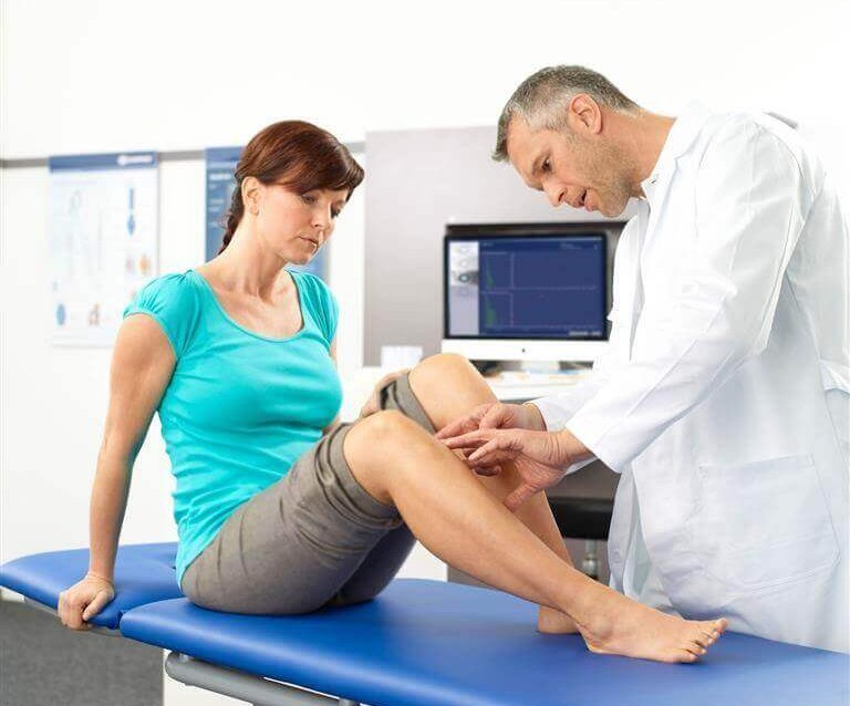Das Bild zeigt eine junge Frau auf einer Liege in einem Behandlungszimmer. Der Arzt untersucht ihre Knie, um die Ursachen sowie eine optimale Behandlung der Knieschmerzen zu identifizieren.